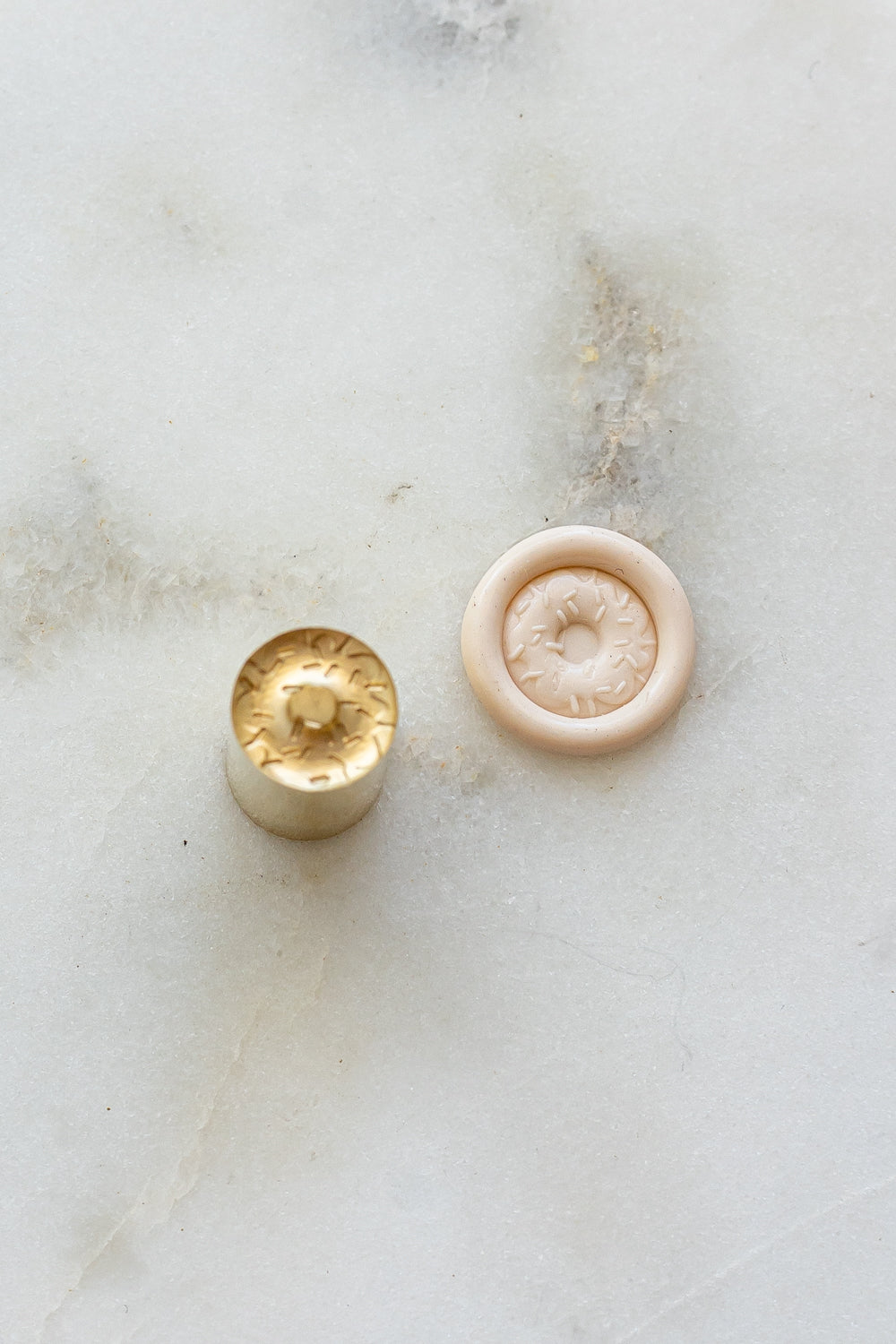 A mini brass wax seal stamp head with a 3D donut design beside a blush wax seal impression on a white marble surface