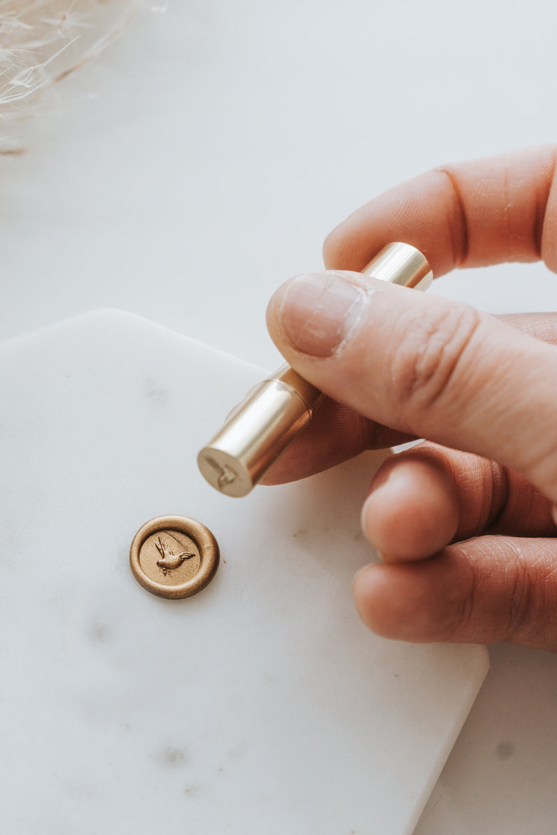 Hand holding brass mini wax seal stamp head with dove design, pressed into gold wax on marble surface