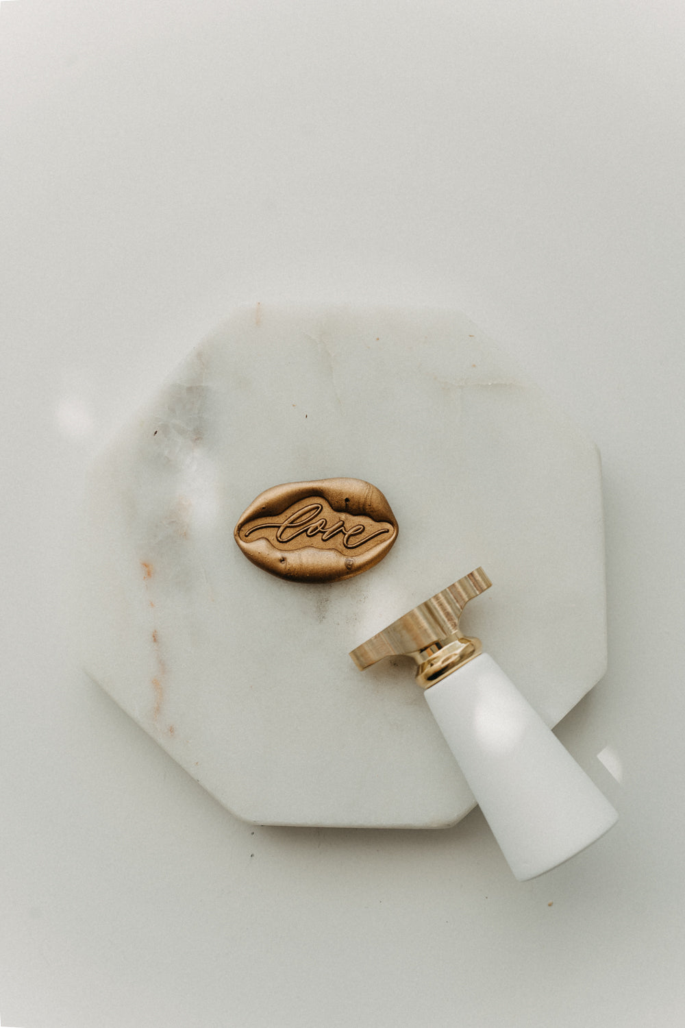 Gold “love” wax seal and white-handled brass wax seal stamp displayed on a white marble tile.