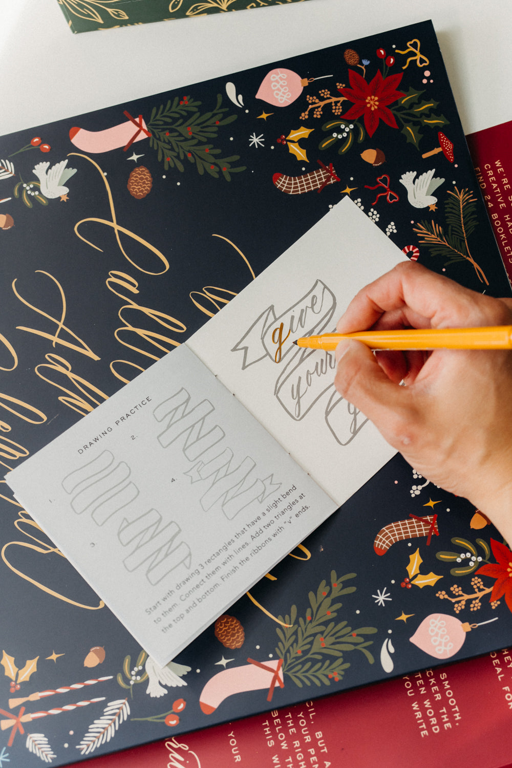 Hand using a yellow brush pen to practice hand lettering in a Christmas calligraphy workbook.