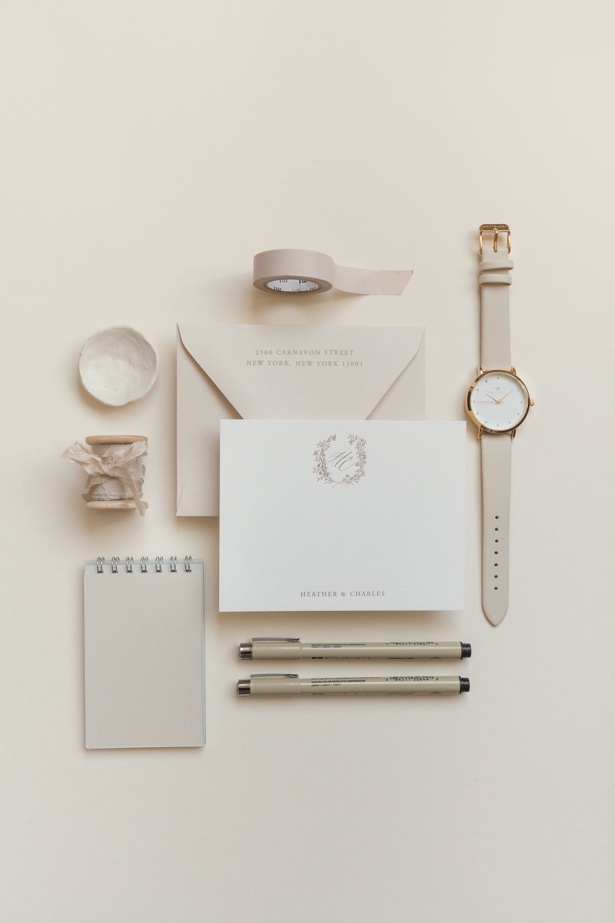 Flat lay wedding stationery with envelope, monogram card, pens, washi tape, and watch in neutral aesthetic