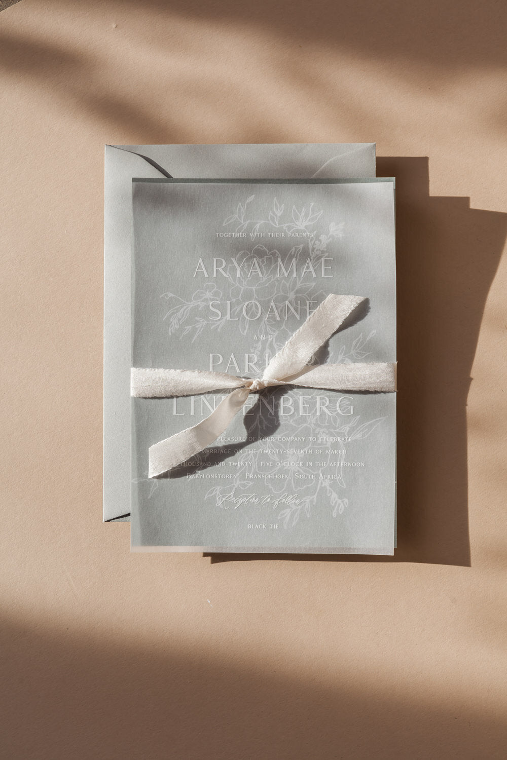 Elegant vellum wedding invitation with white ink floral design, tied with silk ribbon on warm neutral background