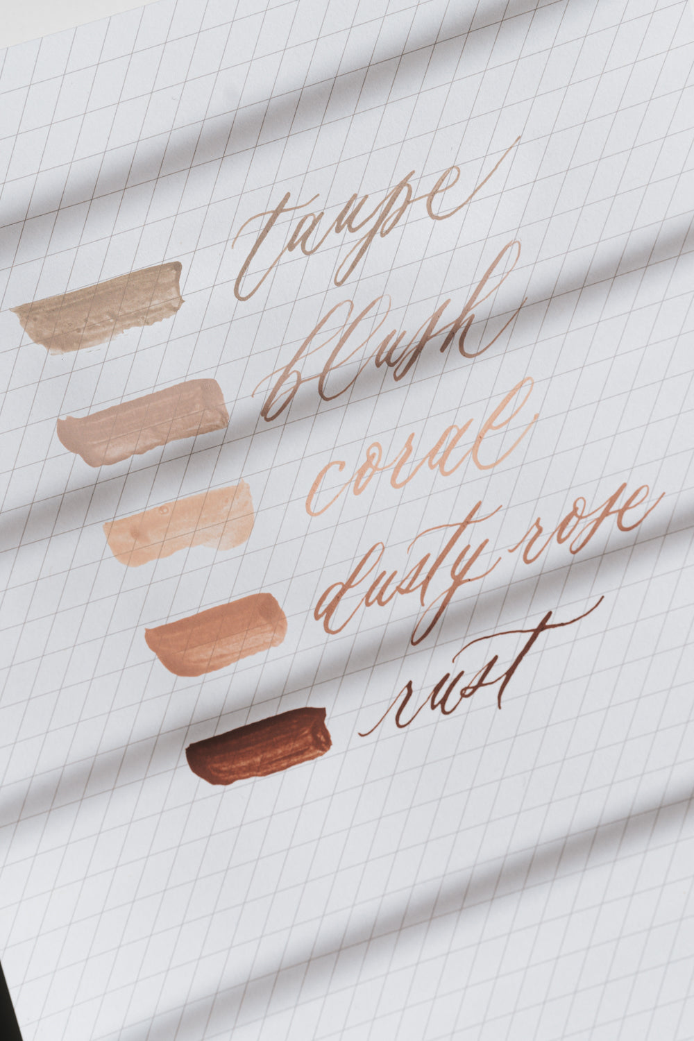 Calligraphy ink swatches labeled taupe, blush, coral, dusty rose, and rust on slanted guide paper in natural light