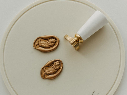 Two gold otter wax seals on a beige silicon mat with its wax stamp beside them