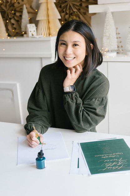 Woman smiling while practicing calligraphy with oblique pen and ink at desk with worksheets and stationery