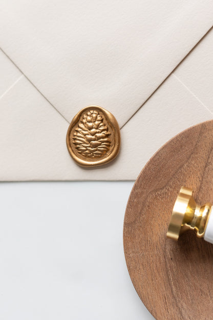 A pinecone wax seal stamp and beige envelope showcasing a gold pinecone wax seal, perfect for Holiday greeting cards
