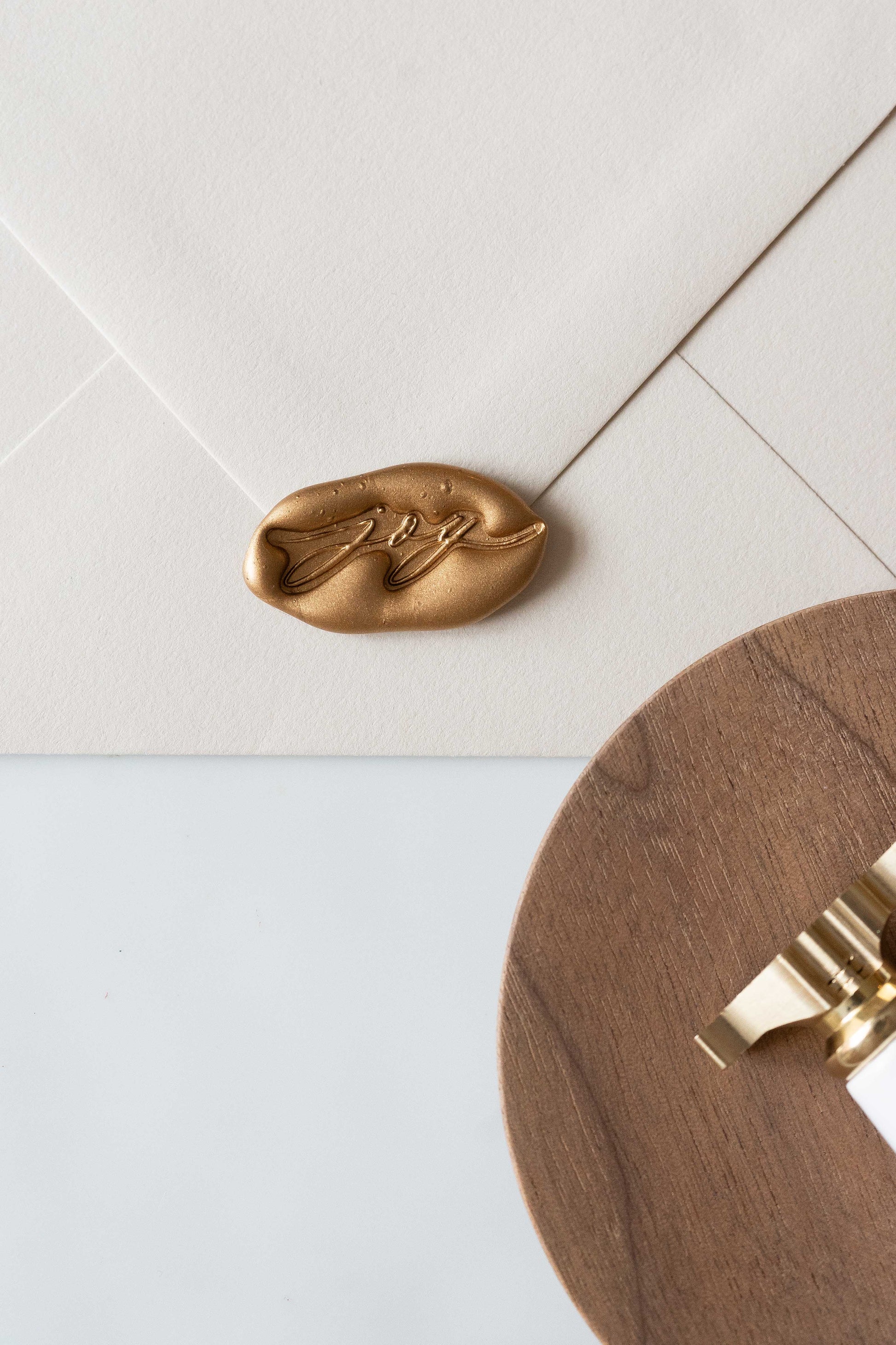 A beige envelope featuring a golden wax seal with the word joy spelled out, perfect for Holiday greeting cards