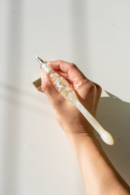 Top view of hand holding ivory resin calligraphy pen with gold flakes on desk, modern calligraphy tools