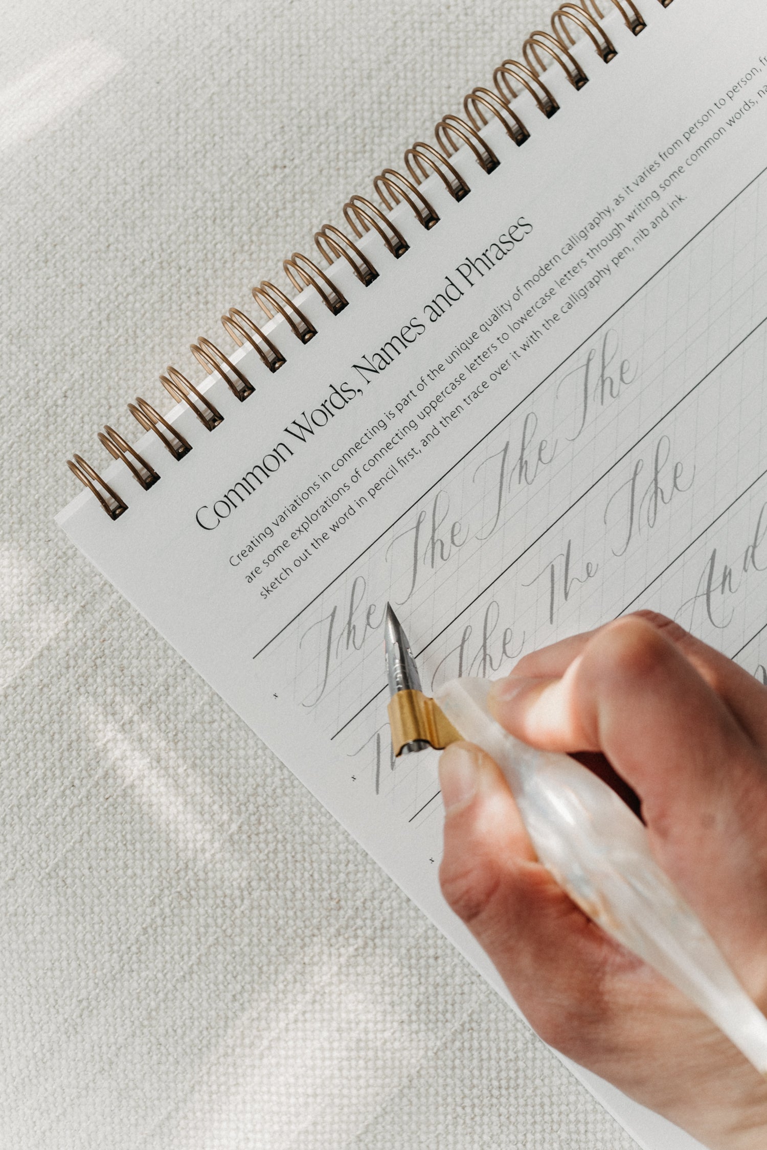 Close-up of a hand holding an oblique pen, tracing calligraphy practice words on a spiral-bound pad titled 'Common Words, Names, and Phrases'
