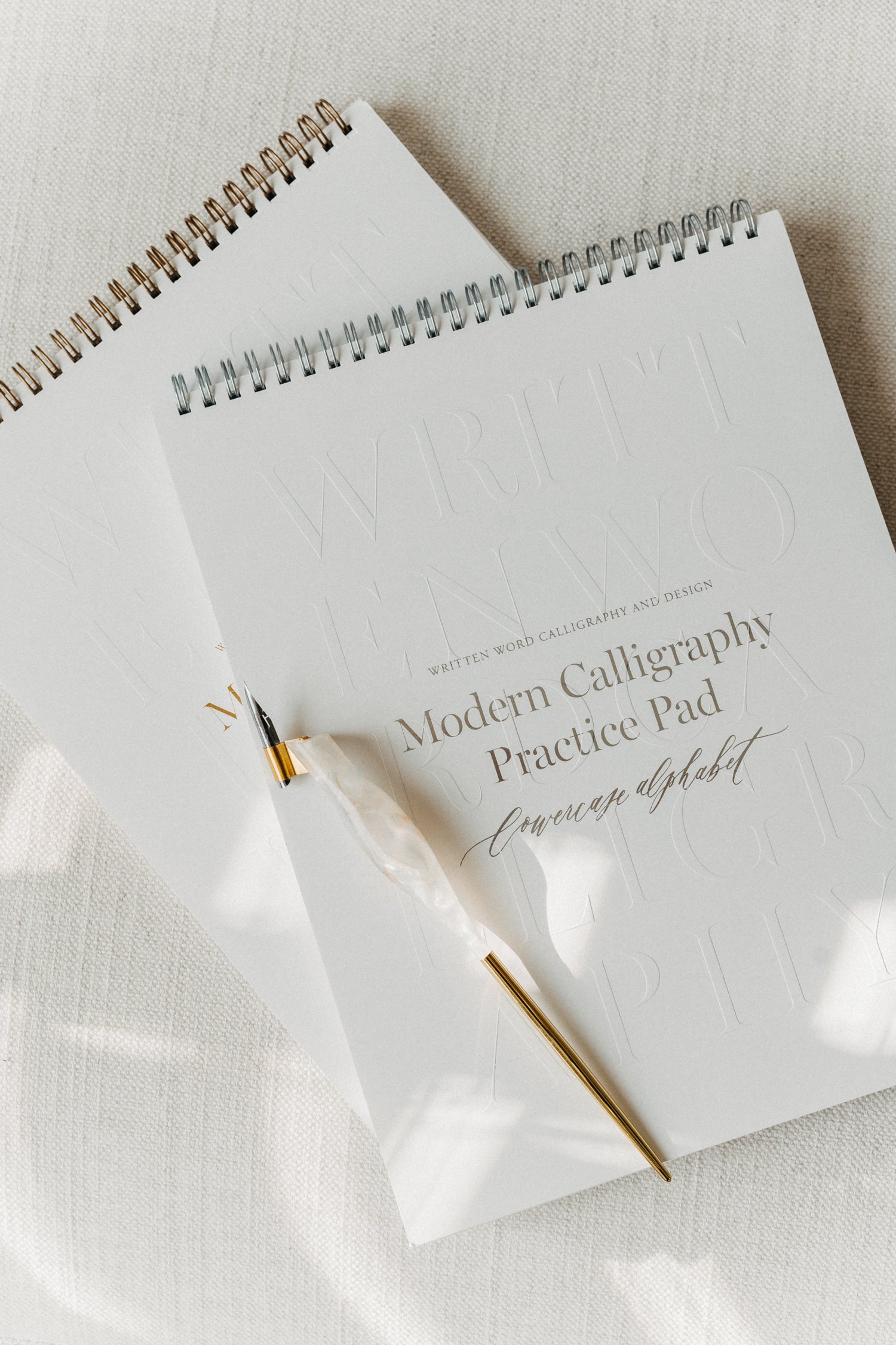 Two modern calligraphy practice pads stacked on a light fabric surface, featuring a quartz pen with a brass flange and nib