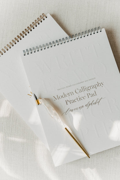 Two modern calligraphy practice pads stacked on a light fabric surface, featuring a quartz pen with a brass flange and nib