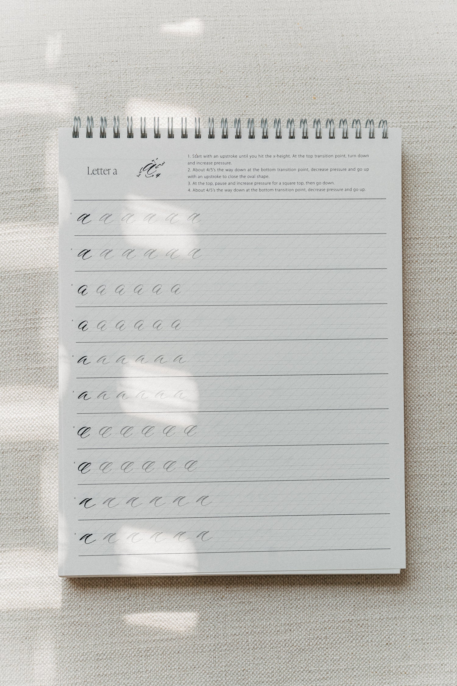 Calligraphy practice pad open to the letter 'a' with detailed instructions and rows of practice strokes on a textured background