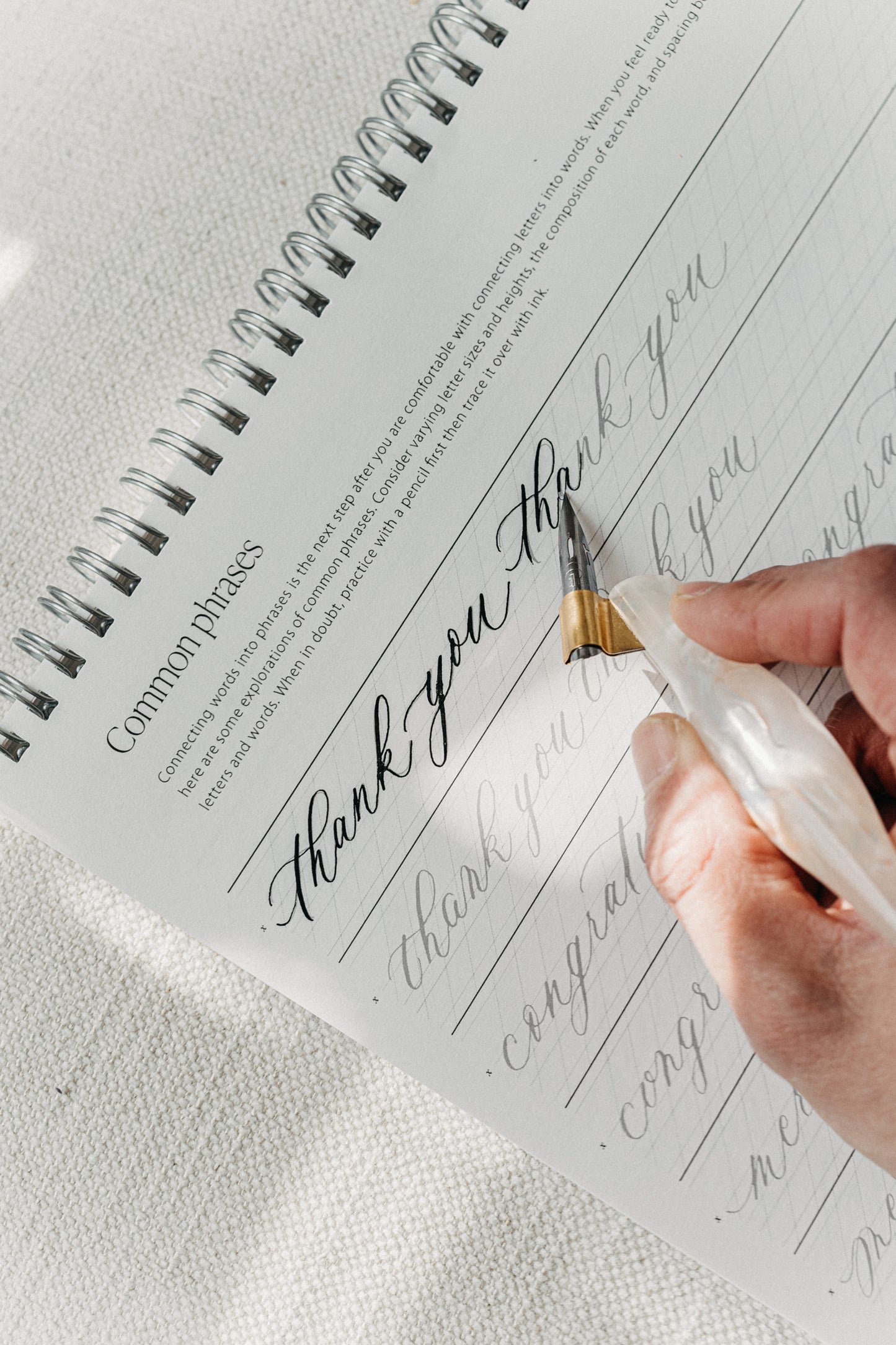 Hand practicing calligraphy of 'Thank you' on a spiral-bound practice pad with a pointed pen, focusing on lettering and spacing