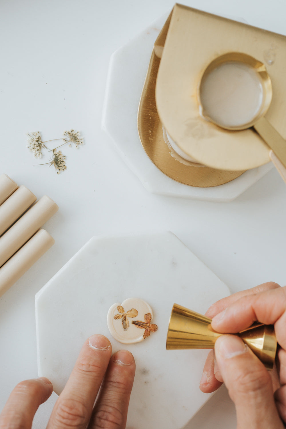A hand pressing a brass wax seal stamp onto melted wax with dried flowers, alongside sealing wax sticks and a wax seal stove.