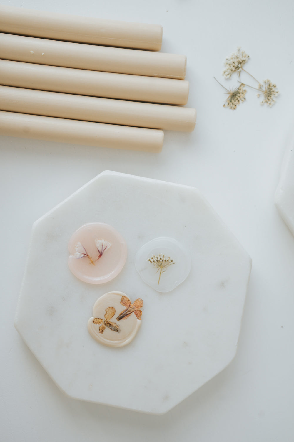 Three handmade wax seals with embedded dried flowers on a white marble tray, next to beige sealing wax sticks and small floral sprigs.