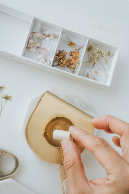A hand melts a sealing wax stick over a gold wax melting spoon, with a box of dried flowers and petals nearby for wax seal embellishments.