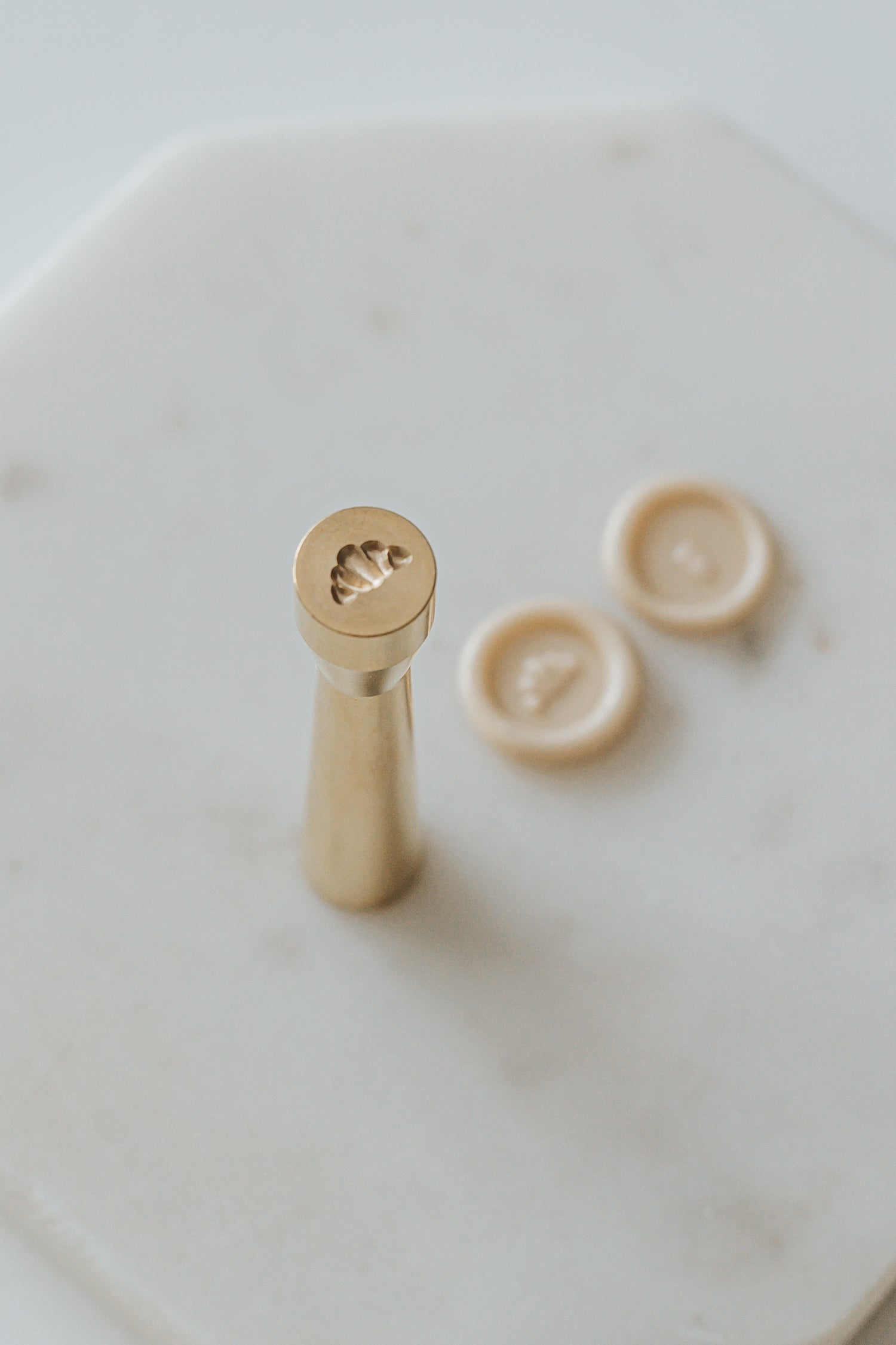 Gold miniature wax seal stamp with croissant designs, placed on a white marble surface with matching beige wax seal