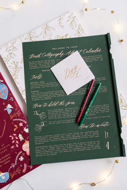 Open Brush Calligraphy Advent Calendar showing a green instruction panel with gold foil text, “Day 1” booklet, and two brush pens in red and green surrounded by warm fairy lights.