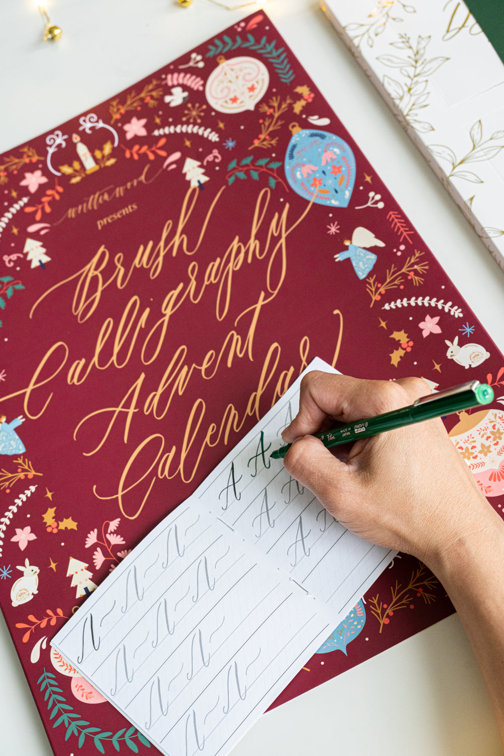 Hand practicing brush lettering on a tracing booklet from the Brush Calligraphy Advent Calendar, placed over the festive maroon box with gold foil lettering and holiday illustrations.