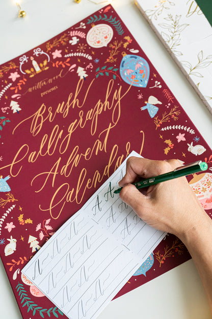 Hand practicing brush lettering on a tracing booklet from the Brush Calligraphy Advent Calendar, placed over the festive maroon box with gold foil lettering and holiday illustrations.