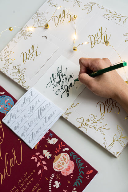 Hand opening a Brush Calligraphy Advent Calendar and practicing the phrase “Abide in Me and I in you” from John 15:4, with gold-foiled doors, twinkle lights, and a maroon illustrated cover.