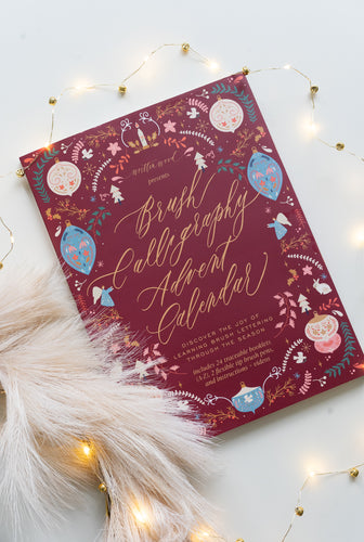 Flatlay of the Brush Calligraphy Advent Calendar with a festive maroon cover, gold foil lettering, and illustrated ornaments, surrounded by fairy lights and soft pampas grass.