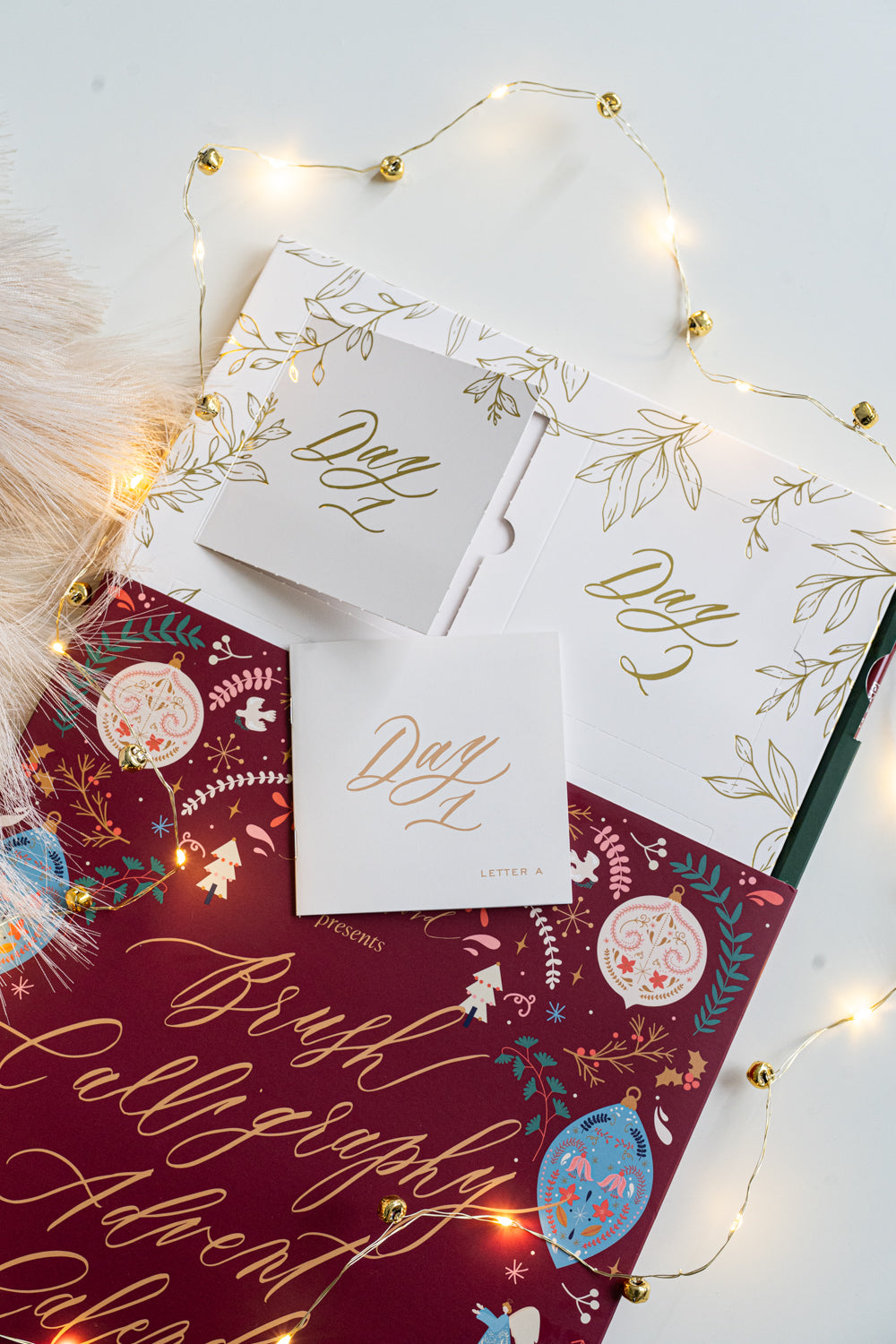 Brush Calligraphy Advent Calendar opened to reveal the “Day 1” and “Day 2” gold foil doors, with the Day 1 booklet displayed on top of the festive maroon cover surrounded by twinkling fairy lights.