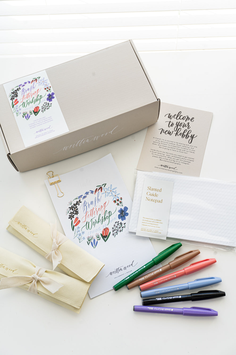 A brush lettering starter kit with a variety of items laid out, including a box with a floral design, brush pens, worksheets, and a notepad, all arranged on a white surface.