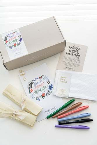 A brush lettering starter kit with a variety of items laid out, including a box with a floral design, brush pens, worksheets, and a notepad, all arranged on a white surface.