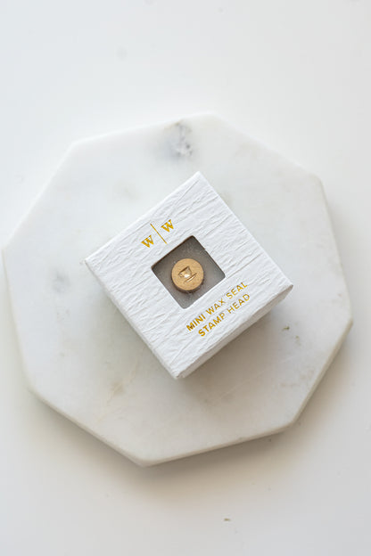 A mini brass wax seal stamp head with a 3D tea cup design in a textured white ring-style box on a marble coaster