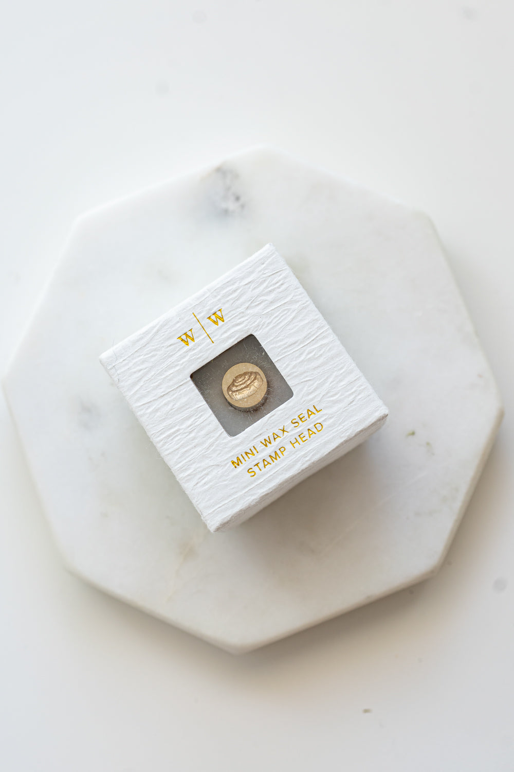 A mini brass wax seal stamp head with a 3D cinnamon bun design in a textured white ring-style box on a marble coaster