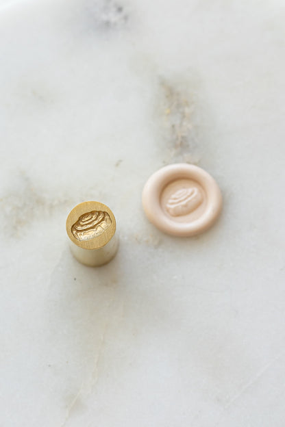​A mini brass wax seal stamp head with a 3D cinnamon bun design beside a blush wax seal impression on a white marble surface