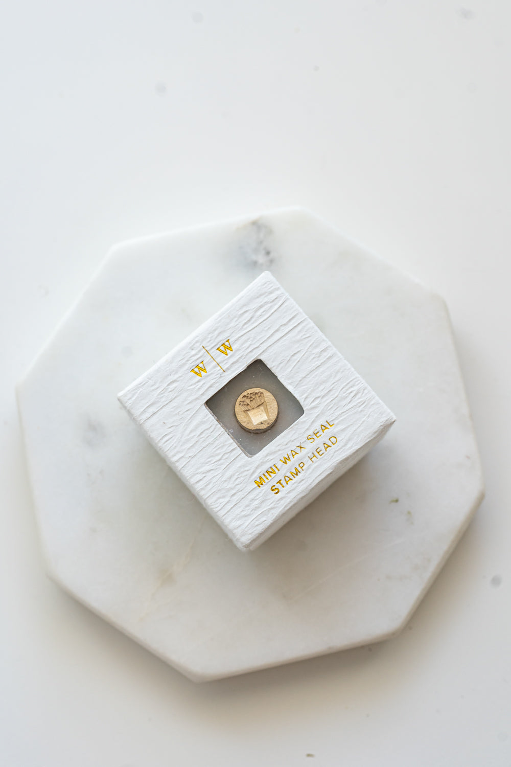A mini brass wax seal stamp head with a 3D French fries design in a textured white ring-style box on a marble coaster