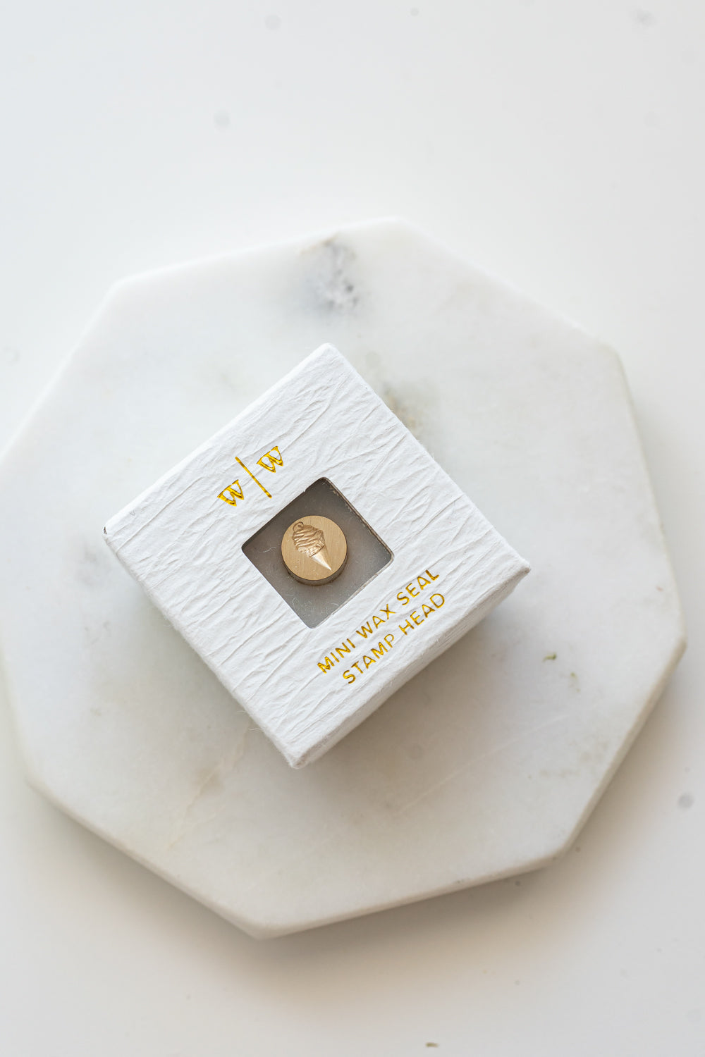 A mini brass wax seal stamp head with a 3D c design in a textured white ring-style box on a marble coaster