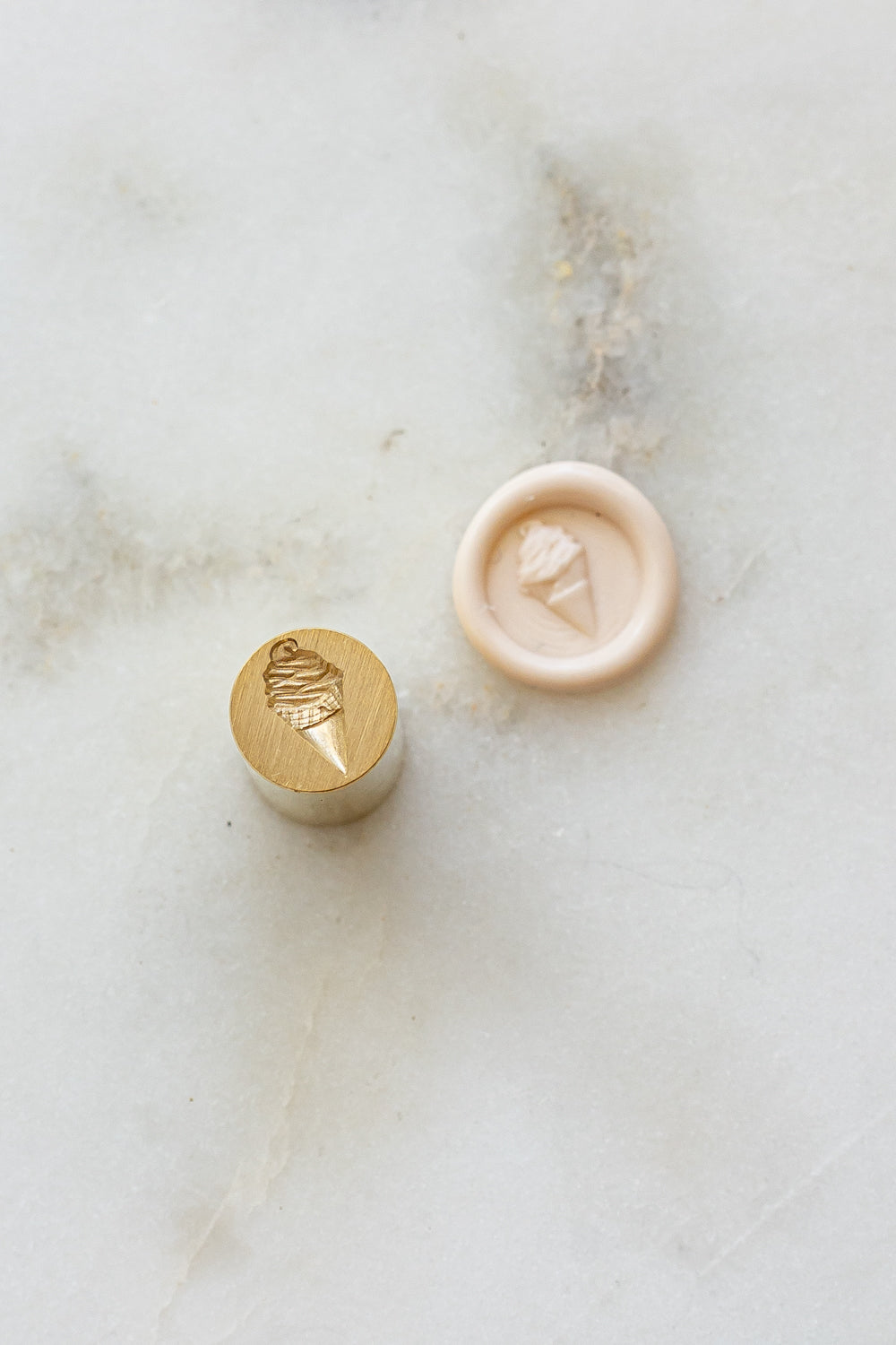 ​A mini brass wax seal stamp head with a 3D soft serve ice cream design beside a blush wax seal impression on a white marble surface