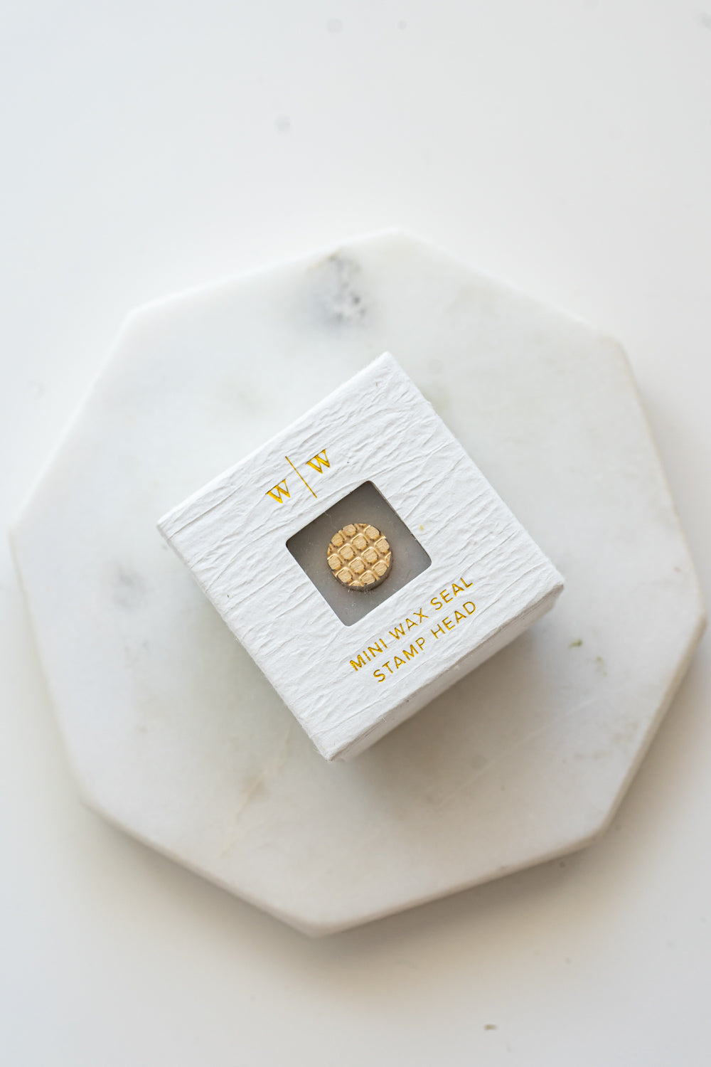 A mini brass wax seal stamp head with a 3D Belgian waffle design in a textured white ring-style box on a marble coaster