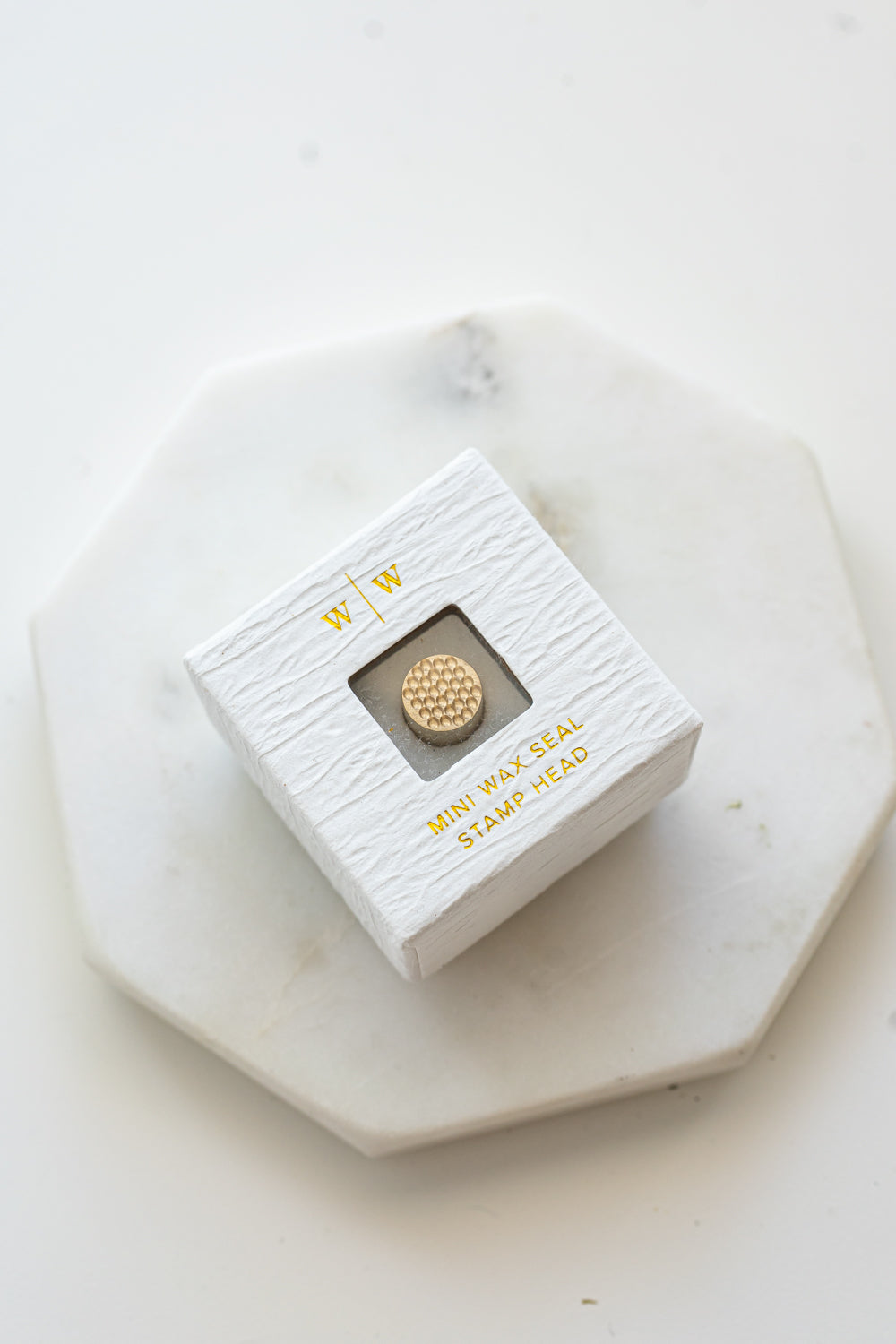 A mini brass wax seal stamp head with a 3D bubble waffle design in a textured white ring-style box on a marble coaster