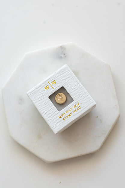 A mini brass wax seal stamp head with a 3D balloons design in a textured white ring-style box on a marble coaster
