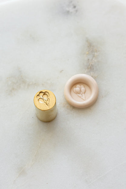 ​A mini brass wax seal stamp head with a 3D baloons design beside a blush wax seal impression on a white marble surface