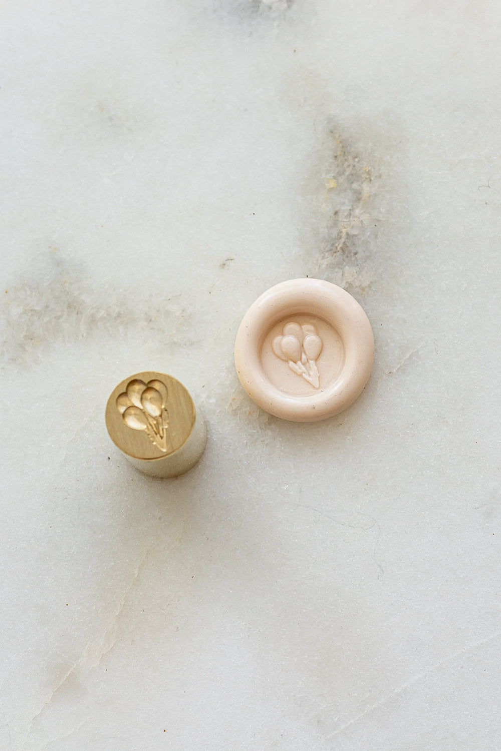 ​A mini brass wax seal stamp head with a 3D balloons design beside a blush wax seal impression on a white marble surface