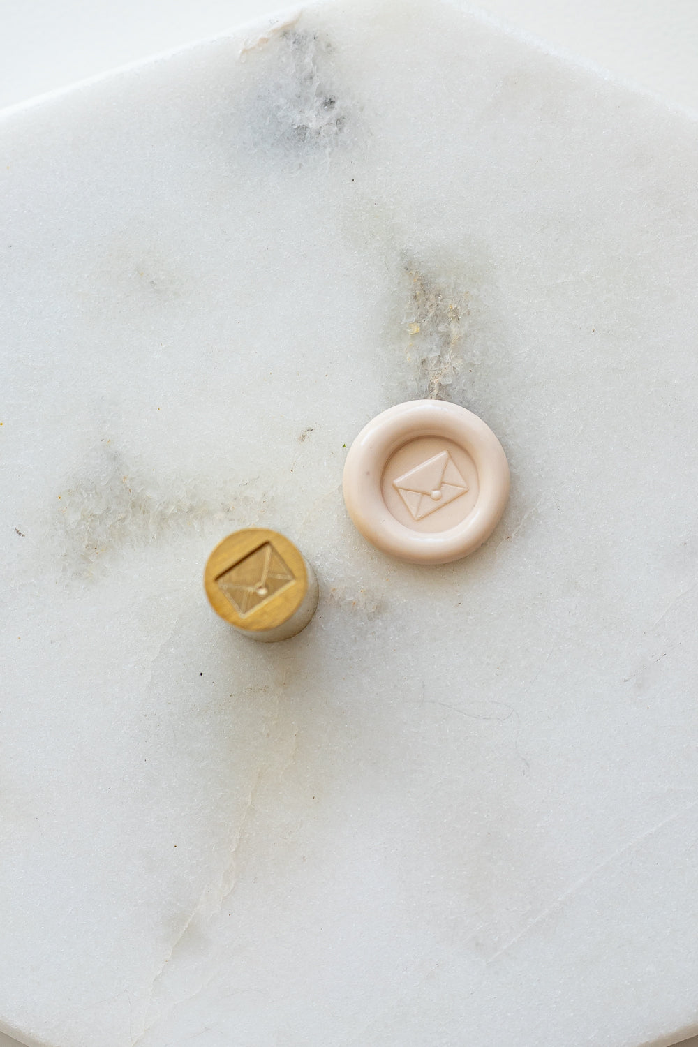 ​A mini brass wax seal stamp head with an envelope design beside a blush wax seal impression on a white marble surface