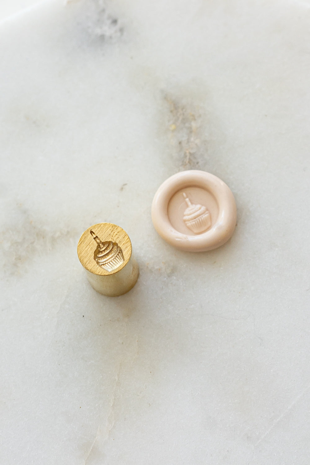​A mini brass wax seal stamp head with a 3D cupcake design beside a blush wax seal impression on a white marble surface
