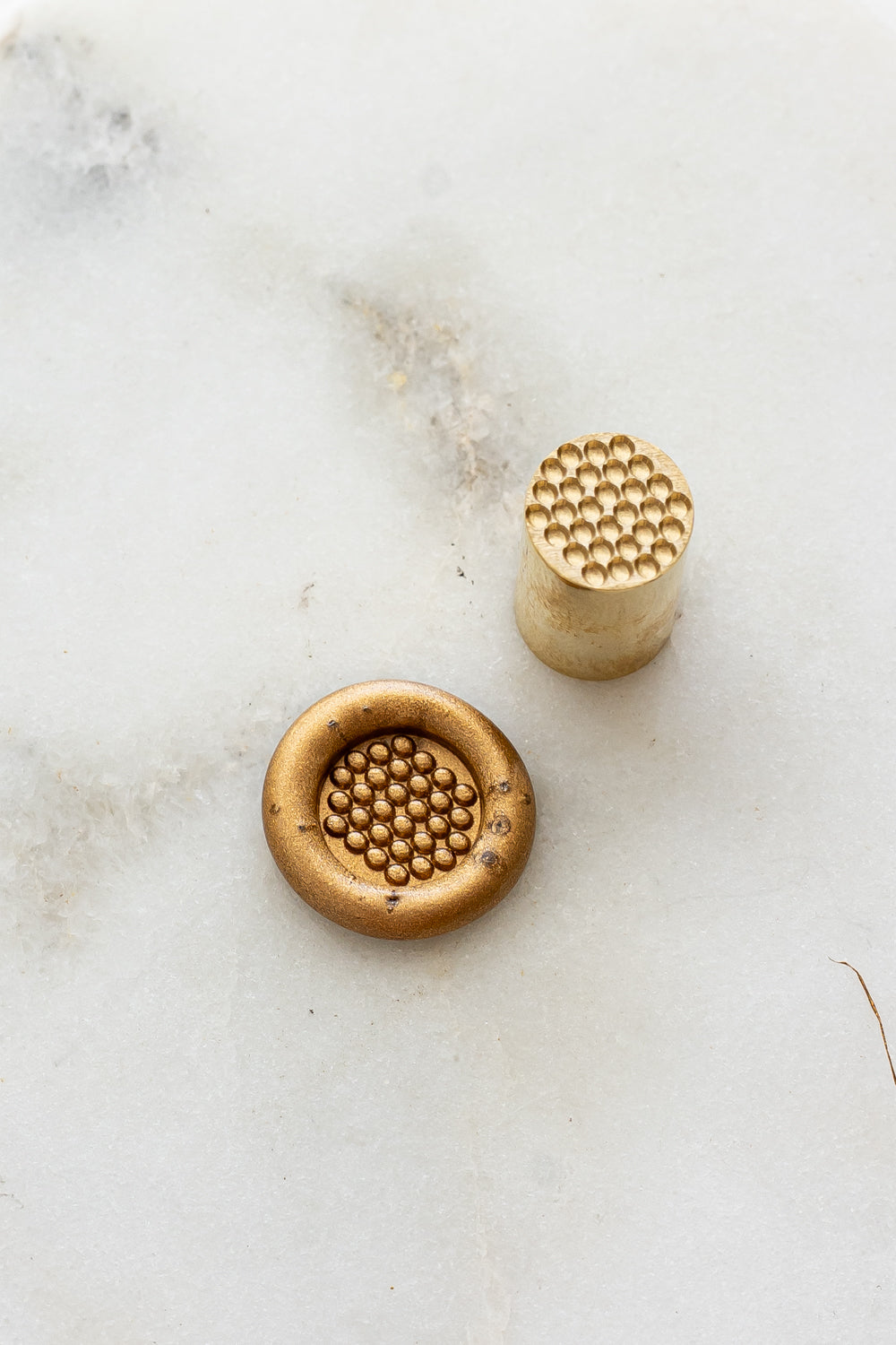 ​A mini brass wax seal stamp head with a 3D bubble waffle design beside a gold wax seal impression on a white marble surface