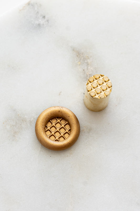 ​A mini brass wax seal stamp head with a 3D Belgian waffle design beside a gold wax seal impression on a white marble surface