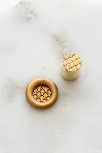 ​A mini brass wax seal stamp head with a 3D Belgian waffle design beside a gold wax seal impression on a white marble surface