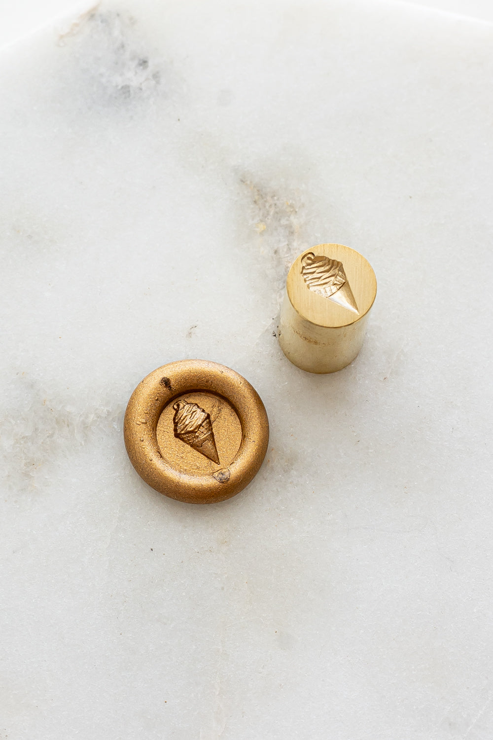 ​A mini brass wax seal stamp head with a 3D soft serve ice cream design beside a gold wax seal impression on a white marble surface