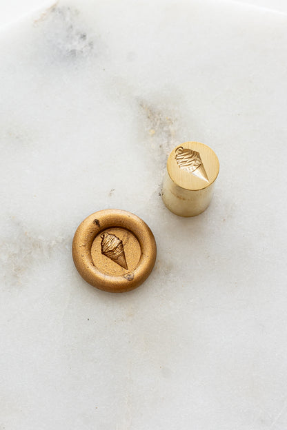 ​A mini brass wax seal stamp head with a 3D soft serve ice cream design beside a gold wax seal impression on a white marble surface
