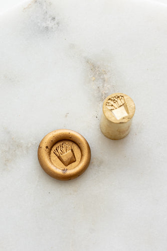 ​A mini brass wax seal stamp head with a 3D French fries design beside a gold wax seal impression on a white marble surface