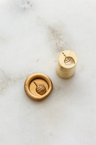​A mini brass wax seal stamp head with a 3D cupcake design beside a gold wax seal impression on a white marble surface
