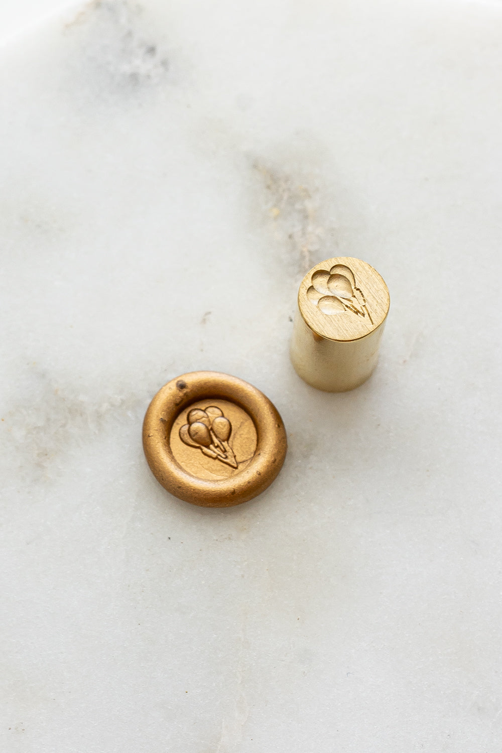 ​A mini brass wax seal stamp head with a 3D balloons design beside a gold wax seal impression on a white marble surface